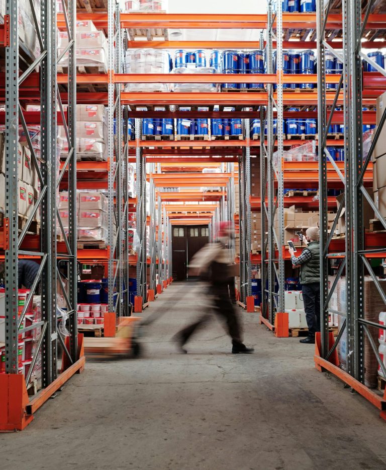 Blurred motion of a man walking in a spacious warehouse aisle between tall shelves.